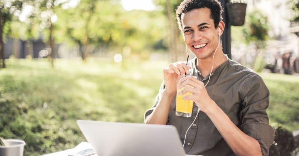 cheerful-guy-with-laptop-and-earphones-sitting-in-park-while-drinking-juice-and-smiling-at-camera