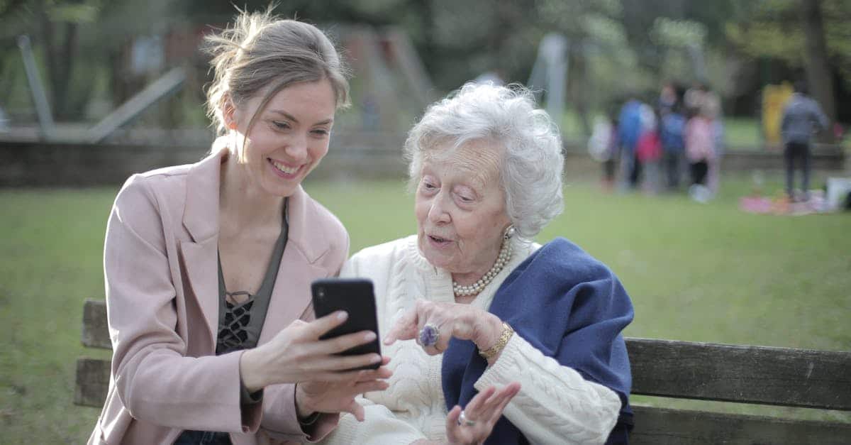 delighted-female-relatives-sitting-together-on-wooden-bench-in-park-and-browsing-mobile-phone-while