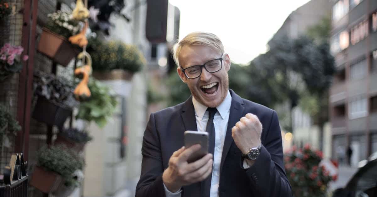 excited-young-man-in-formal-wear-and-eyeglasses-clenching-fist-and-rejoicing-victory-while-using-mob