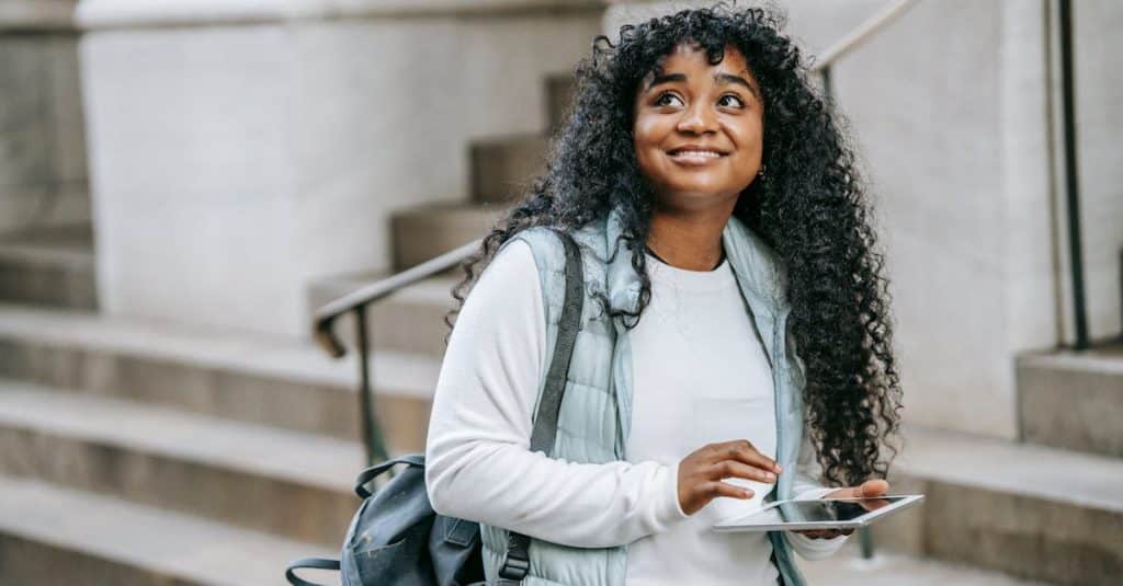 happy-young-african-american-woman-wearing-casual-clothes-with-backpack-while-using-tablet-on-city-s