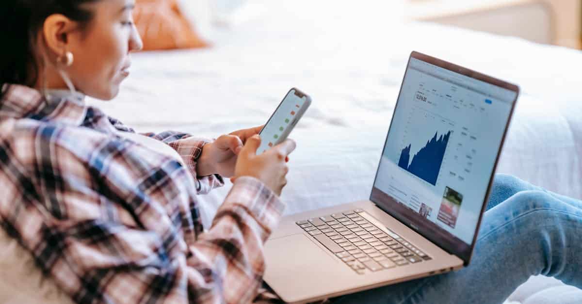 side-view-of-crop-ethnic-female-dealer-using-trading-app-on-cellphone-against-netbook-with-graph-on