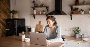 young-female-in-gray-sweater-laughing-while-sitting-at-wooden-table-with-laptop-and-milk-bottle-and